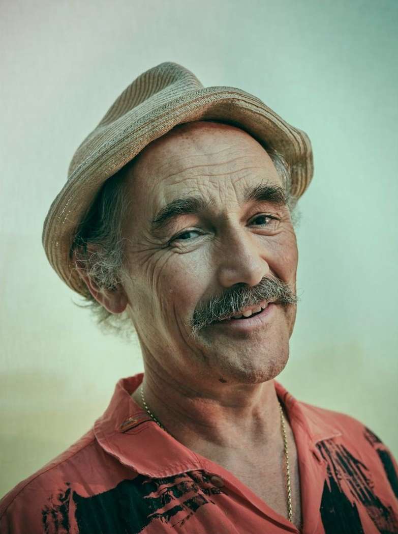Close-up portrait of a smiling, mature man with a mustache wearing a straw hat and coral shirt against a soft, diffused background.