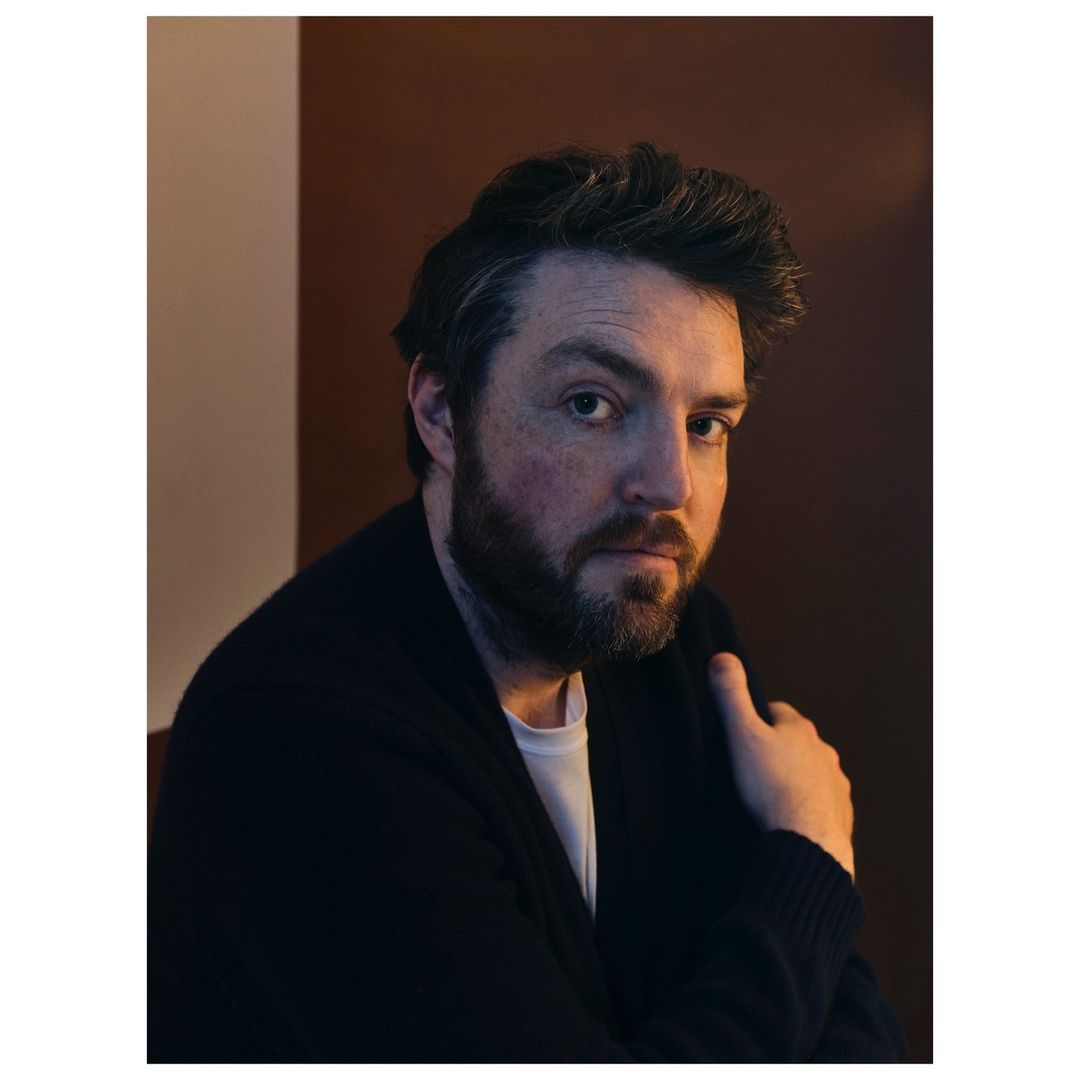 Portrait of a man with a beard wearing a white t-shirt and dark cardigan, posing in a warm, indoor setting with soft, directional lighting.