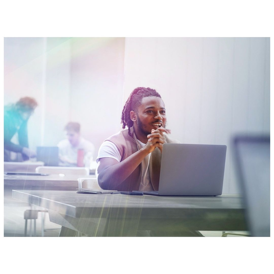 Adult male sits at a table with a laptop in a bright office setting, appearing thoughtful with other people in the background.