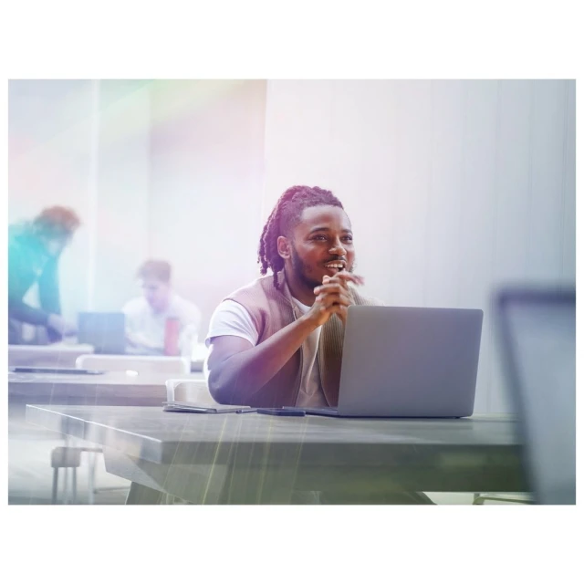 Adult male sits at a table with a laptop in a bright office setting, appearing thoughtful with other people in the background.