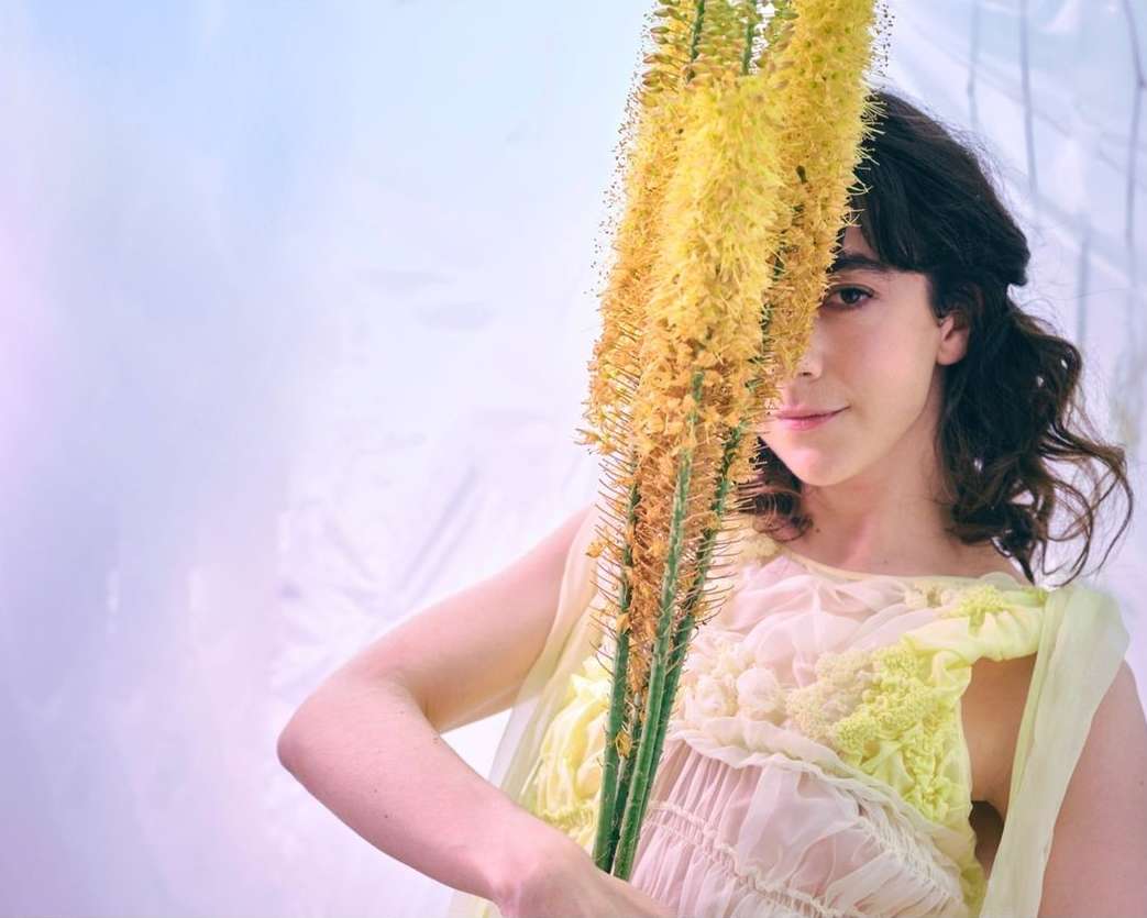 Woman poses indoors with yellow foxtail lilies. The fair-skinned female wears a sheer yellow garment. Soft lighting creates a whimsical, dreamy aesthetic.