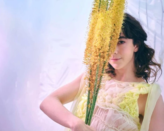Woman poses indoors with yellow foxtail lilies. The fair-skinned female wears a sheer yellow garment. Soft lighting creates a whimsical, dreamy aesthetic.