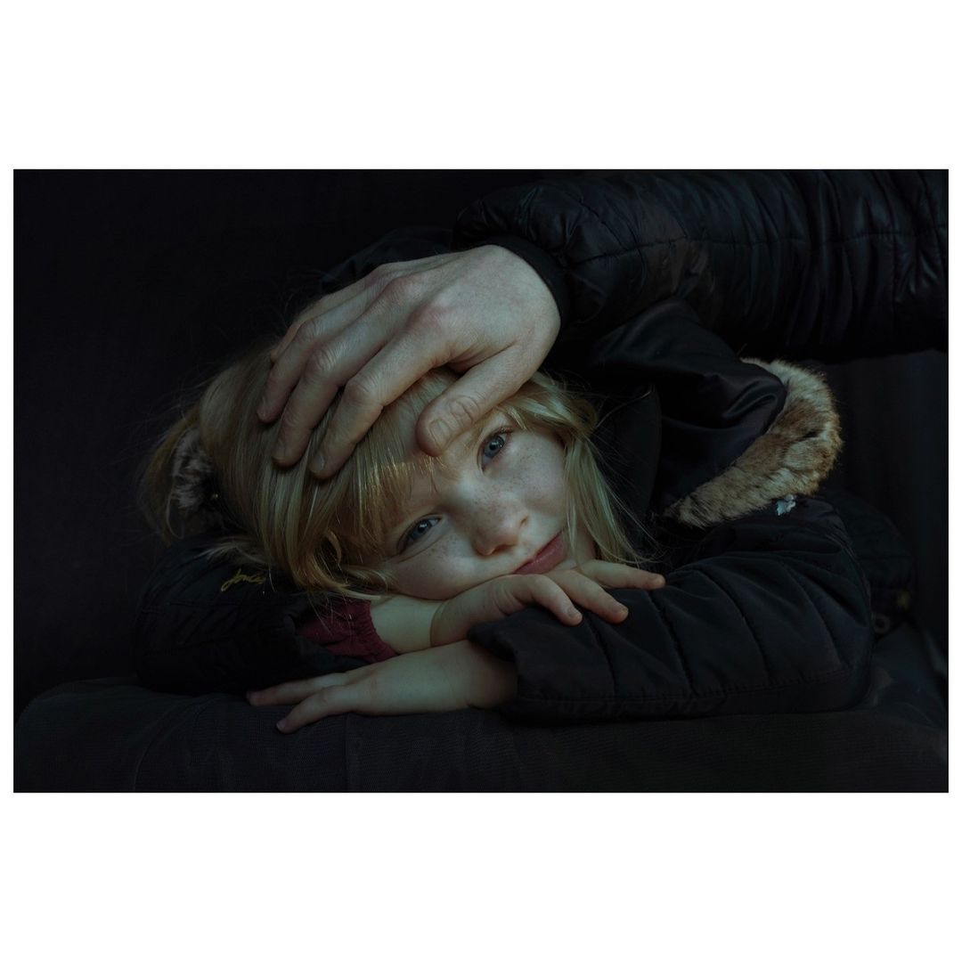A young blonde girl in a dark coat rests her head on her crossed arms while an adult's hand rests protectively on her head in a dark setting.