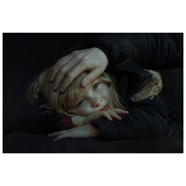 A young blonde girl in a dark coat rests her head on her crossed arms while an adult's hand rests protectively on her head in a dark setting.