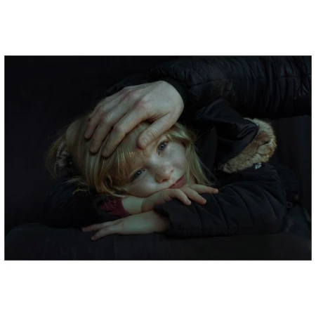 A young blonde girl in a dark coat rests her head on her crossed arms while an adult's hand rests protectively on her head in a dark setting.
