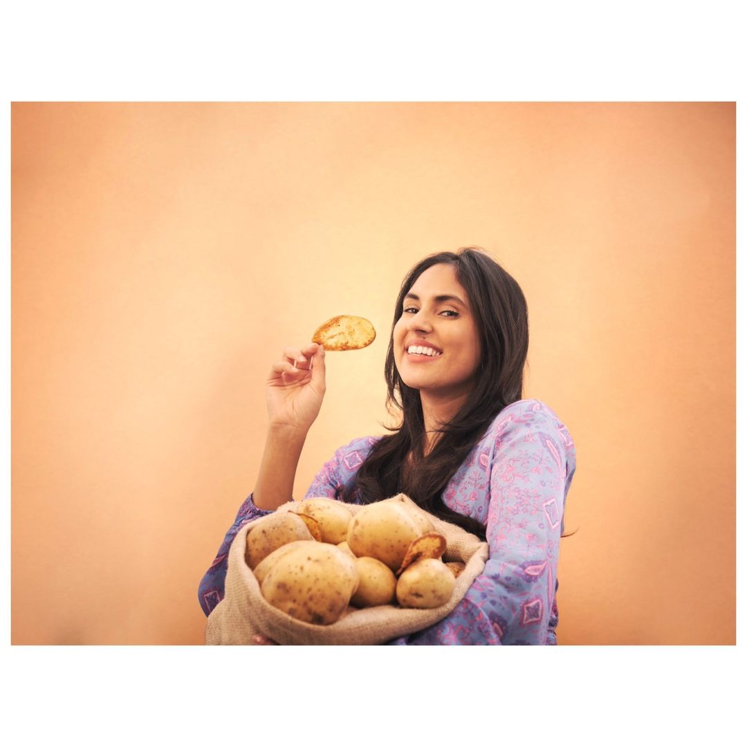 Smiling woman holds potato chip and burlap sack of potatoes against orange background, advertising food or promoting healthy eating.