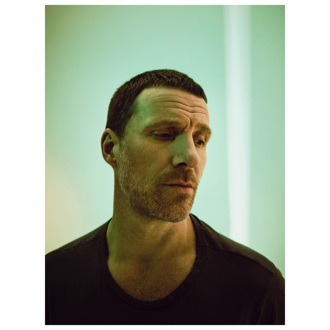 Portrait of a mature adult man wearing a black t-shirt against a soft green background. He has short dark hair and a beard.