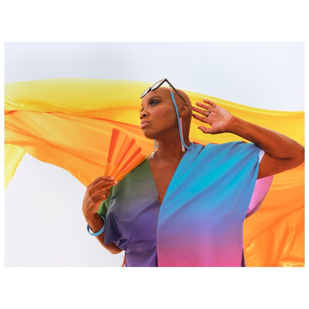 Elegant woman with short hair and dark glasses poses with a rainbow-colored fan and flowing yellow fabric against a white background.