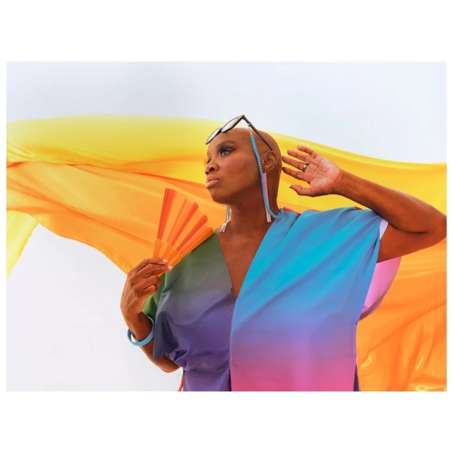 Elegant woman with short hair and dark glasses poses with a rainbow-colored fan and flowing yellow fabric against a white background.