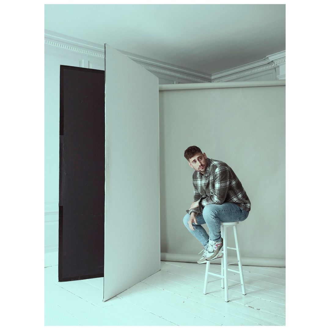 Young man with a beard sits on a white stool in a studio with black and white panels, wearing a plaid shirt and light-wash jeans.