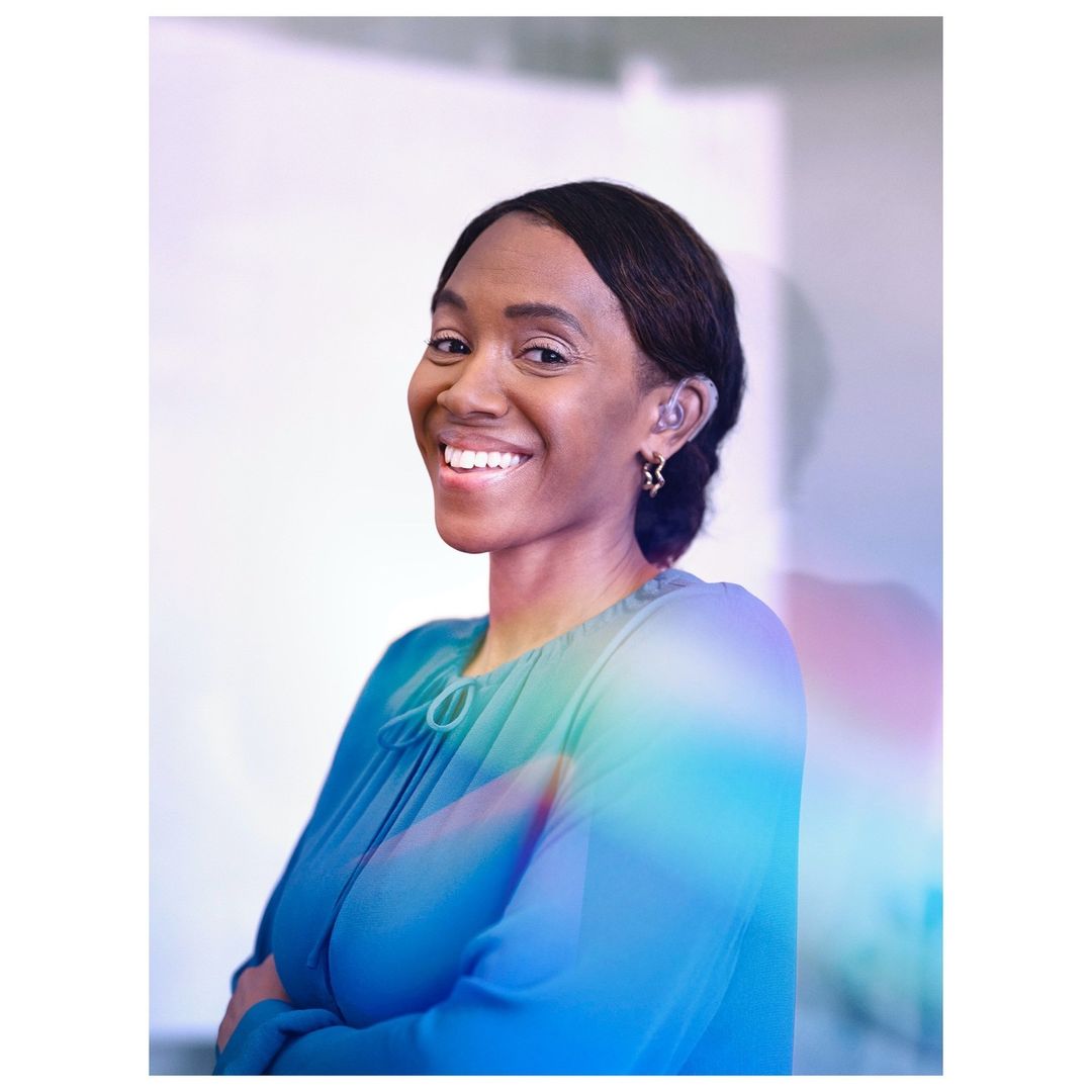Smiling African American woman with hearing aid wearing a blue blouse and star earrings with her arms crossed indoors.