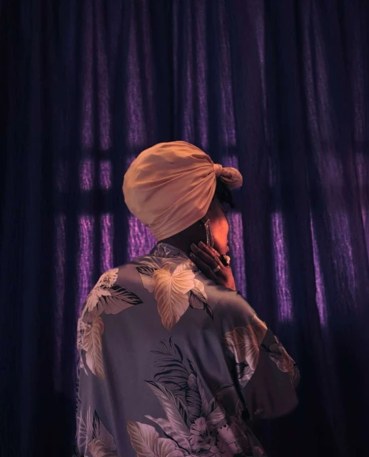 An adult woman is seen from the back wearing a headscarf and robe in a soft, muted color palette against a dark purple backdrop.