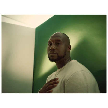 Portrait of a man in a light sweater with hand on chest, posing against a green and grey wall in soft, diffused natural lighting.