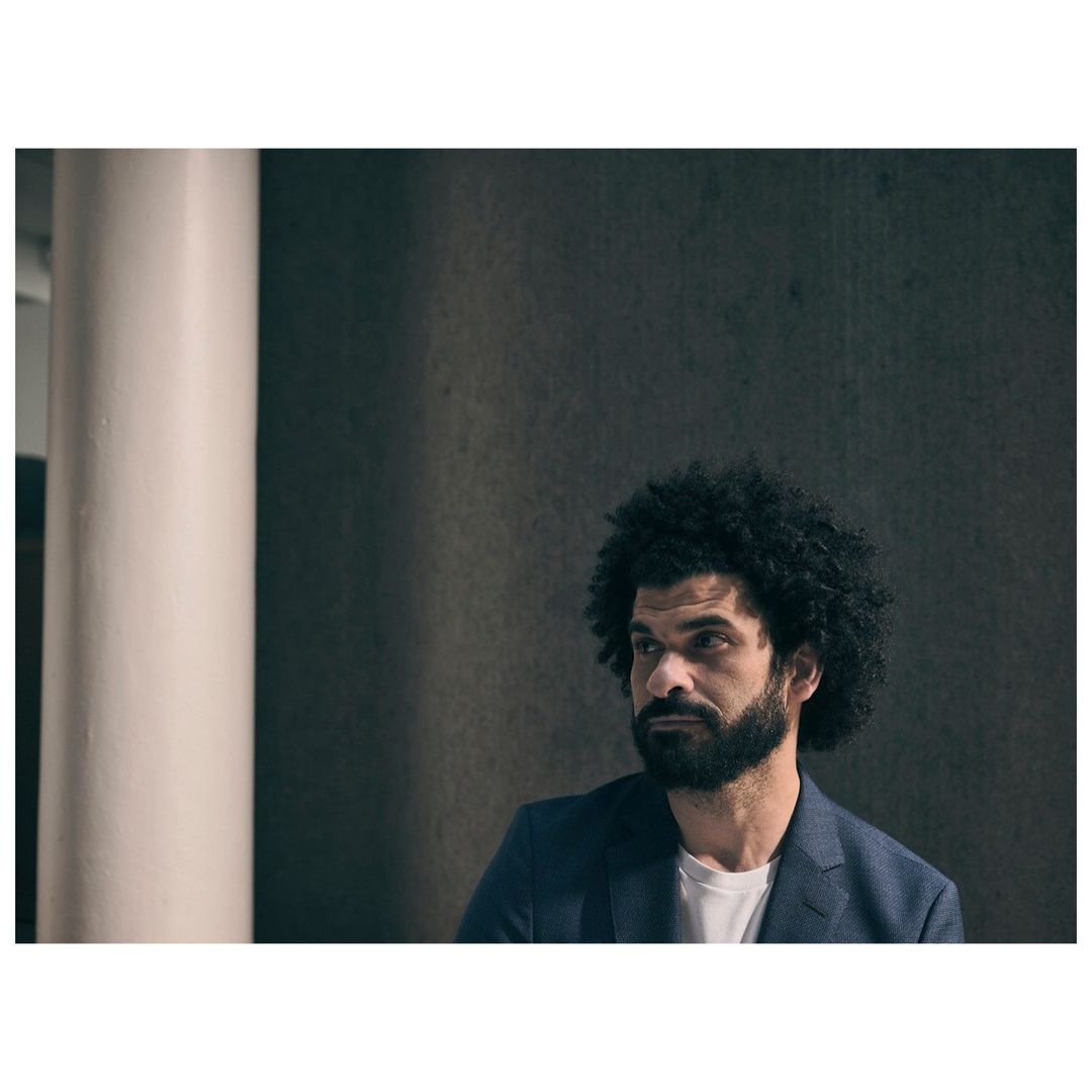 Portrait of a man with dark curly hair and a beard wearing a dark blazer over a white t-shirt, photographed by Phil Fisk.