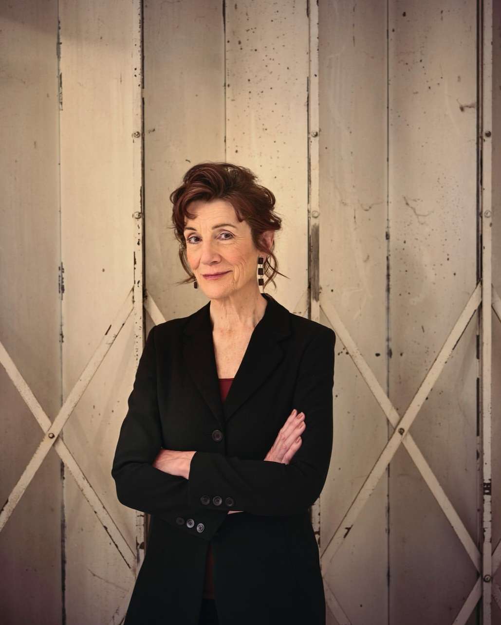 Portrait of an adult woman with short, auburn hair wearing a black jacket, smiling, with arms crossed against a white paneled background.
