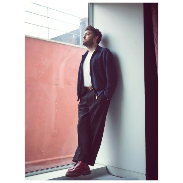 A man in a blue jacket and black pants stands in a corner near a window, looking out with a neutral expression in a naturally lit scene.