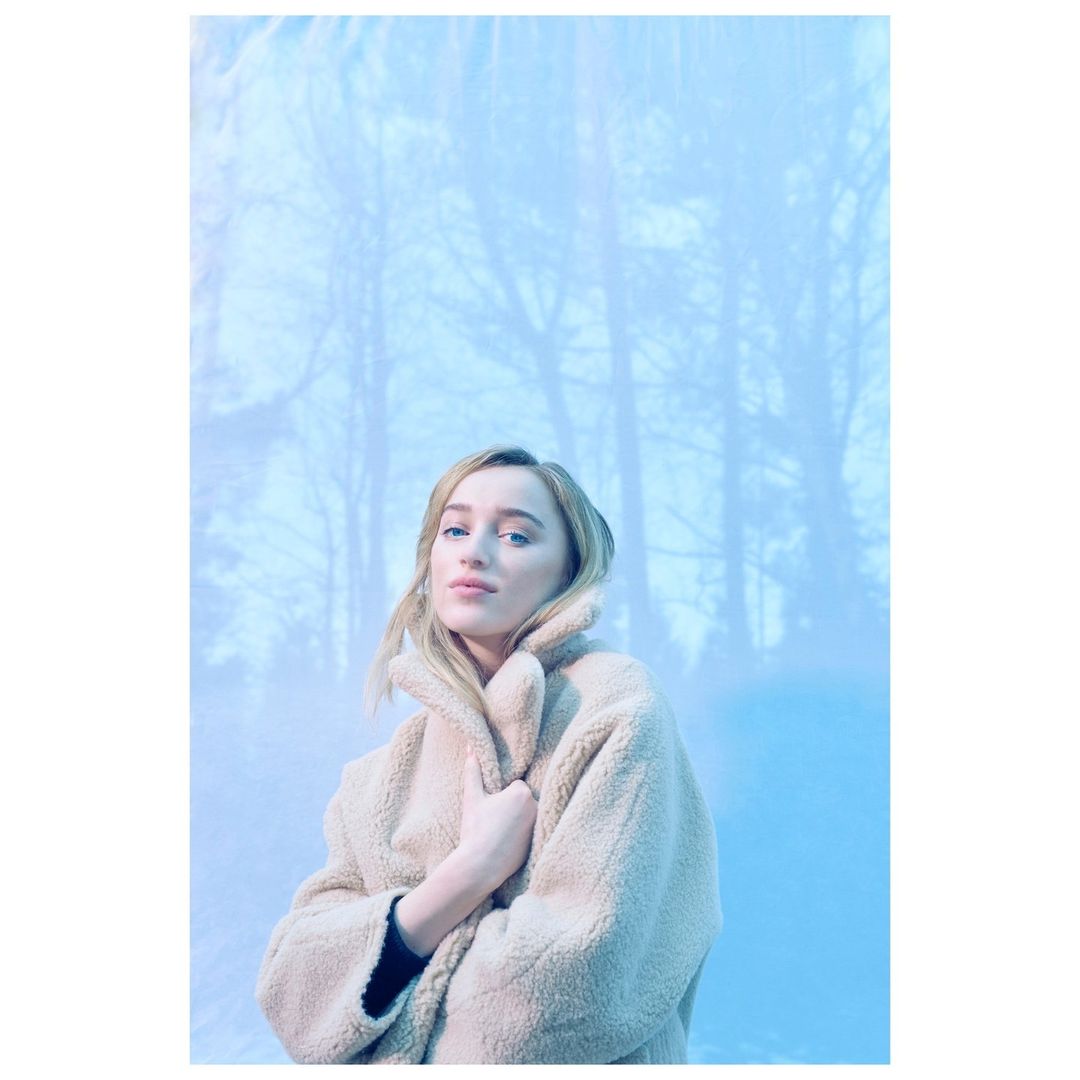 Portrait of an adult female model with blonde hair wearing a tan coat, posing in front of a blue backdrop of a forest in winter.