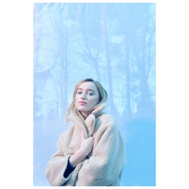 Portrait of an adult female model with blonde hair wearing a tan coat, posing in front of a blue backdrop of a forest in winter.
