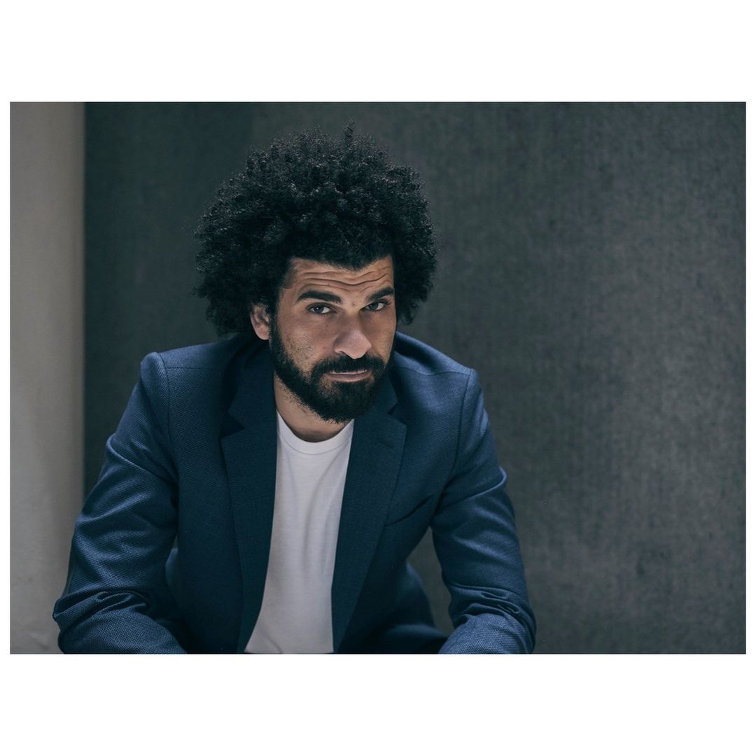 Portrait of a man with curly hair and a beard wearing a dark suit jacket and white t-shirt by photographer Phil Fisk