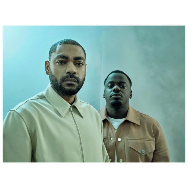 Two men, one with a beard, stand in front of a teal wall. The man in front wears a beige jacket, while the second man wears a tan jacket.