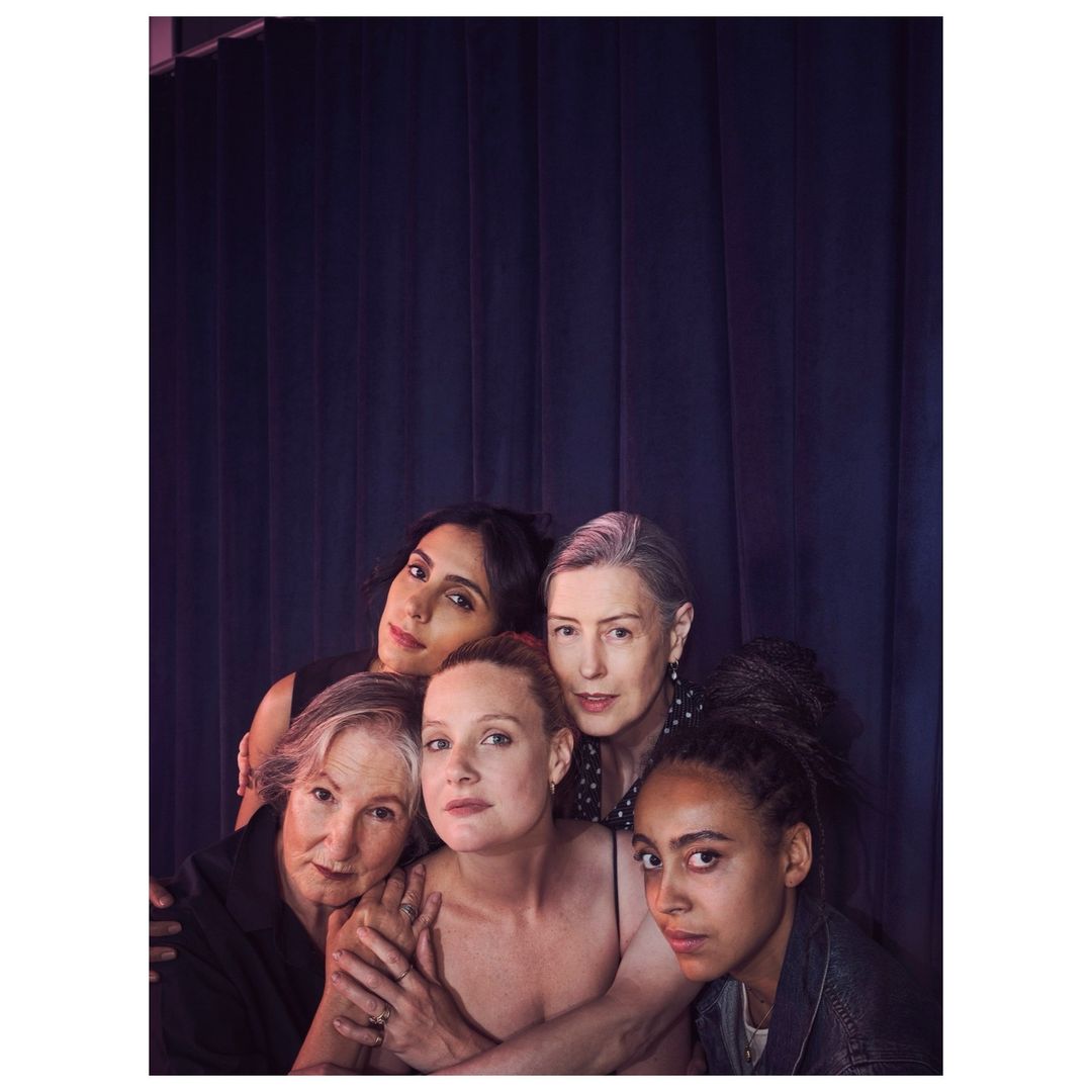 Group portrait of five women of diverse ages and ethnicities, posing closely in front of a dark purple draped backdrop in soft lighting.