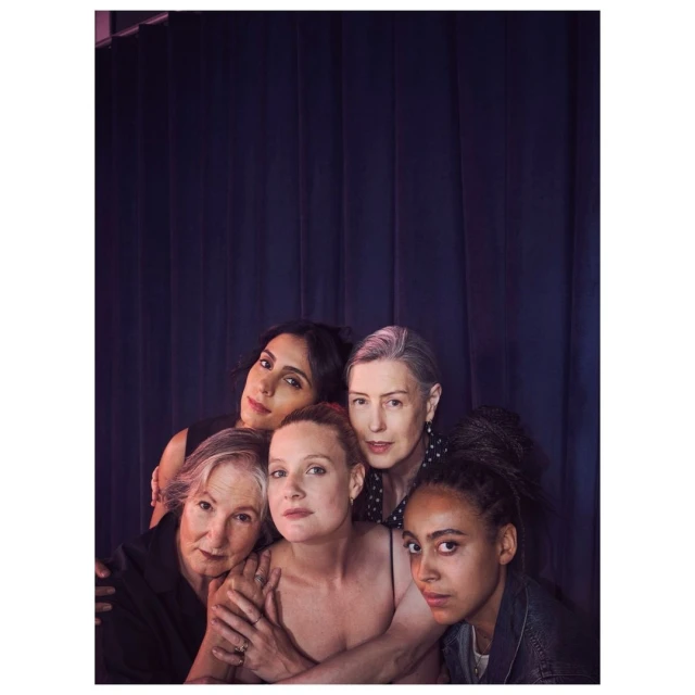 Group portrait of five women of diverse ages and ethnicities, posing closely in front of a dark purple draped backdrop in soft lighting.