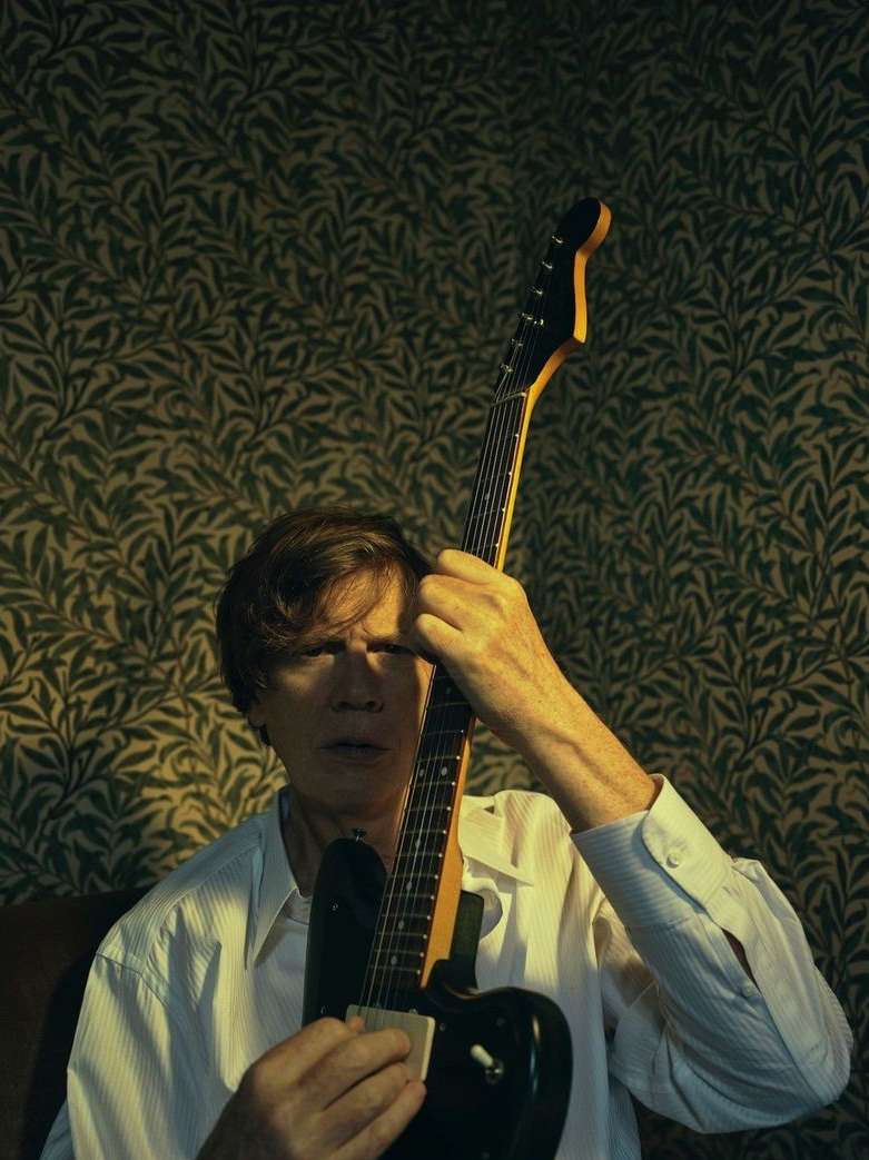 Adult male playing an electric guitar in an indoor setting with patterned wallpaper, wearing a white shirt, medium shot.