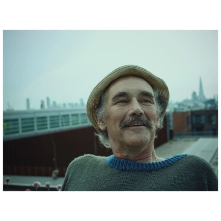 Portrait of an elderly man wearing a straw hat and grey sweater against an outdoor urban skyline backdrop by Phil Fisk.