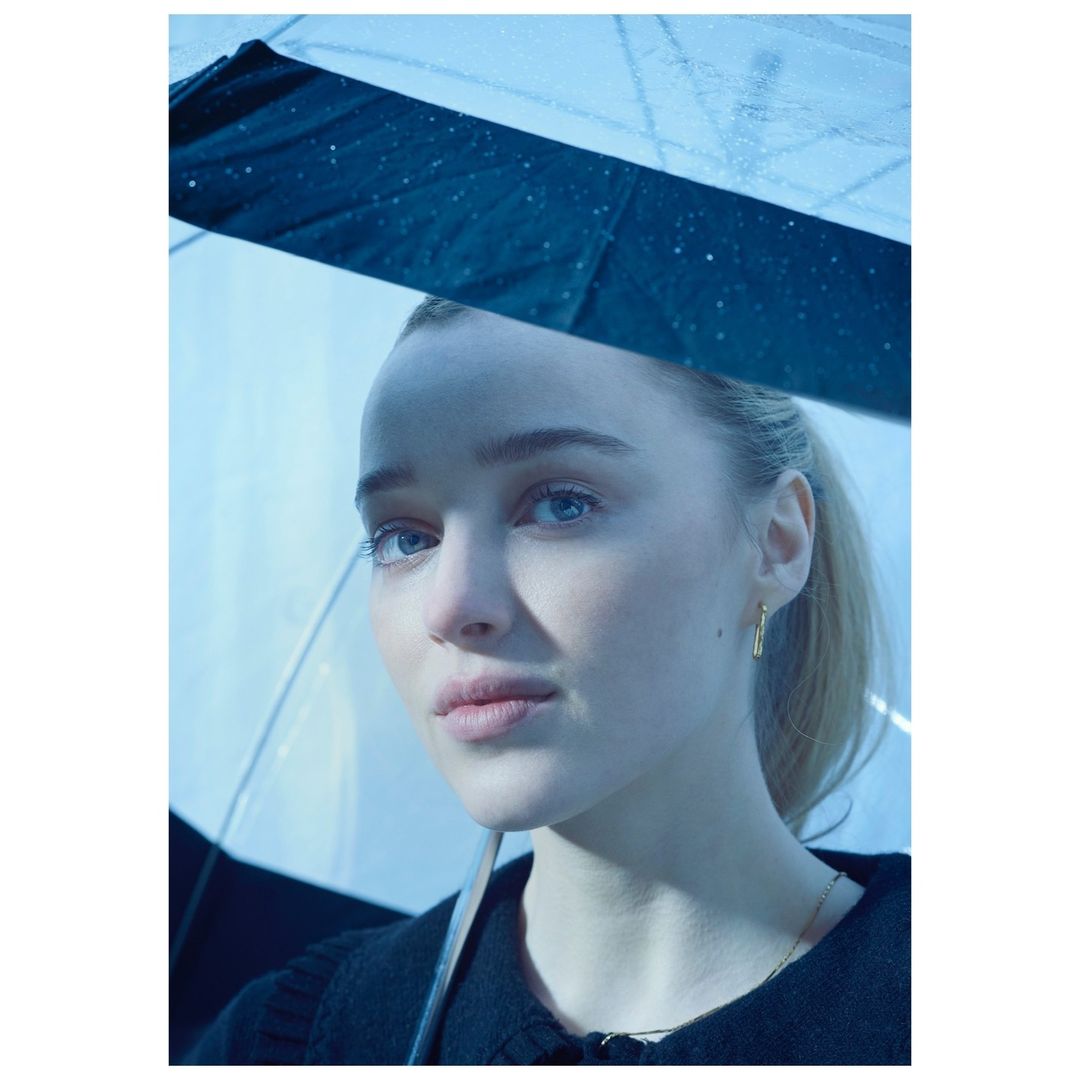 Portrait of a young woman under a transparent umbrella, looking thoughtfully to the side. The lighting is soft and diffused.