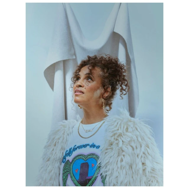 Portrait of an adult woman looking up, wearing a white furry jacket and graphic t-shirt, with a white backdrop in a studio setting.