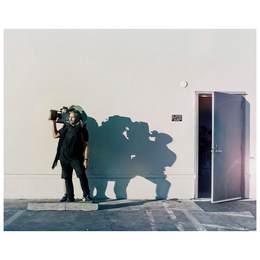 Adult male stands outdoors with a large film camera on his shoulder in front of a building; daytime, sunny.