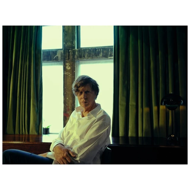 Indoor portrait of a mature adult male in a white button-down shirt sitting near a window with green curtains and a black table lamp.