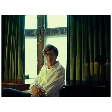 Indoor portrait of a mature adult male in a white button-down shirt sitting near a window with green curtains and a black table lamp.