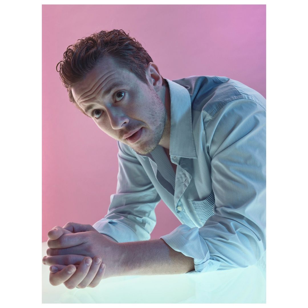 Headshot of a fair-skinned adult male with curly brown hair, wearing a striped dress shirt, leaning forward, with a pink-and-blue background.