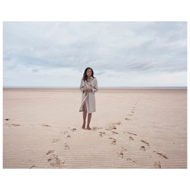 Adult female walking barefoot on a sandy beach wearing a trench coat, beneath a cloudy sky, with footprints in the sand.
