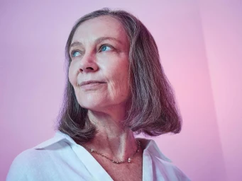Portrait of an elderly woman with short gray hair wearing a white collared shirt against a pink gradient background.