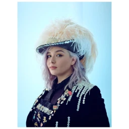 Portrait of a person with pastel purple hair wearing a feathered hat and embellished dark jacket