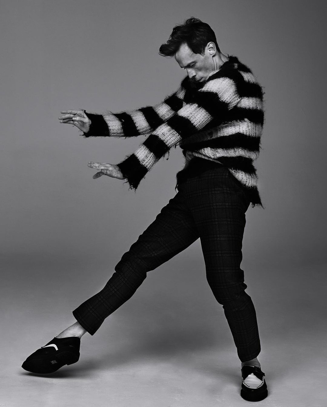 Black and white studio shot of an adult male posing in a striped sweater and plaid pants with studio lighting and a neutral backdrop.