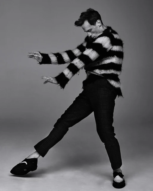 Black and white studio shot of an adult male posing in a striped sweater and plaid pants with studio lighting and a neutral backdrop.