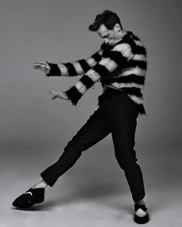 Black and white studio shot of an adult male posing in a striped sweater and plaid pants with studio lighting and a neutral backdrop.