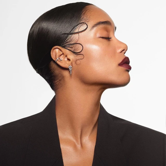 Portrait of a Black woman in a black blazer with slicked-back hair and decorative hair strands, set against a bright white backdrop.