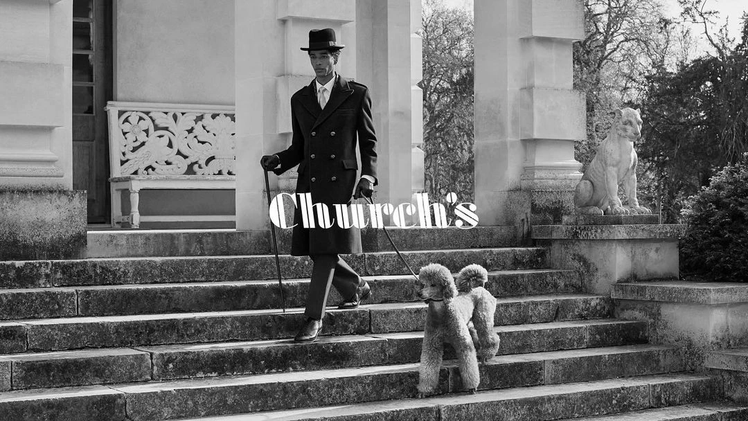 Elegant black and white image of a stylish adult male descending stone steps with two poodles, wearing a coat and hat, and holding a cane.