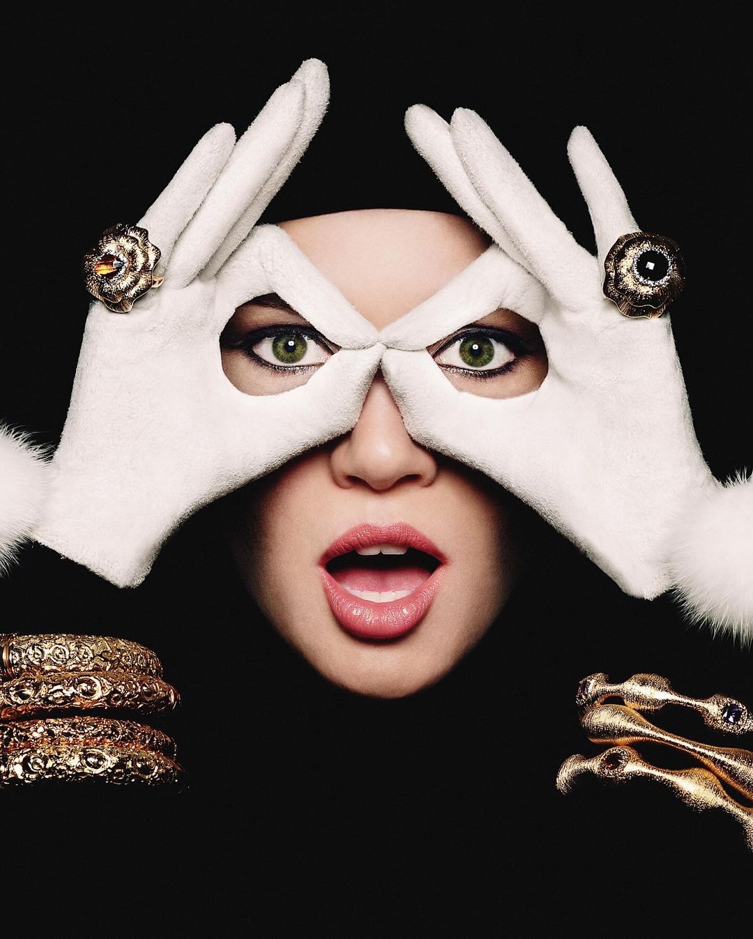Close-up of a woman with surprised expression wearing white gloves and jewelry against a black background in a studio setting.