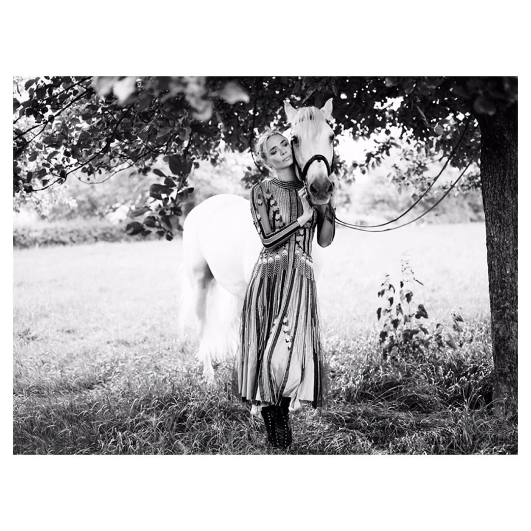 Monochrome shot of a woman in a dress standing next to a white horse in a grassy field, underneath a tree with leaves, creating a serene scene.