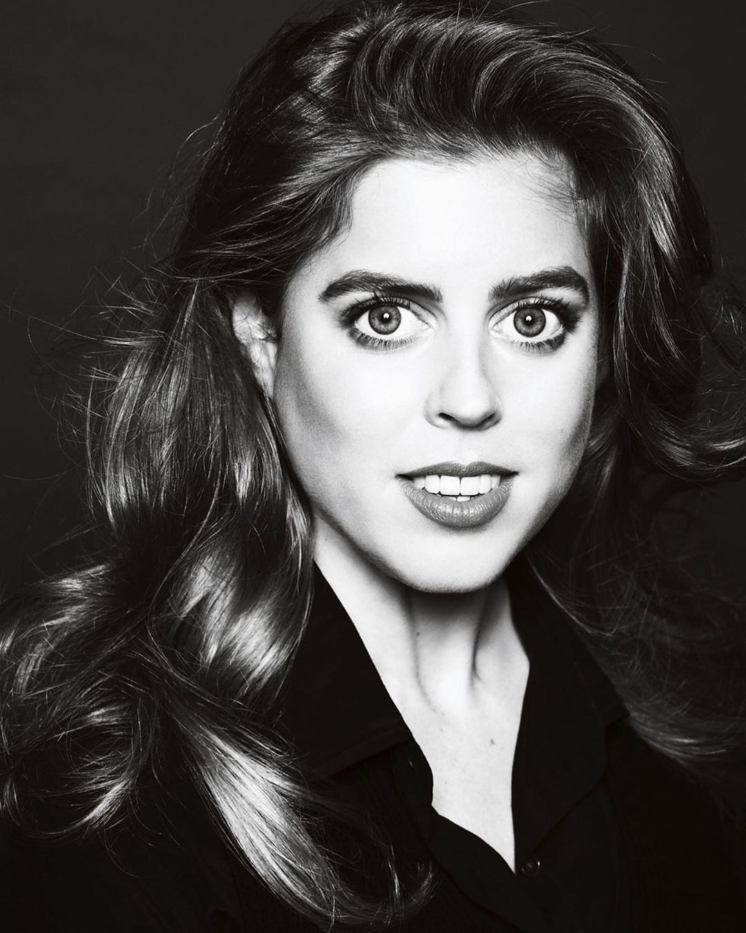 Headshot of a light-skinned adult woman with long wavy brown hair wearing a dark shirt, looking directly at the camera in a photo studio.