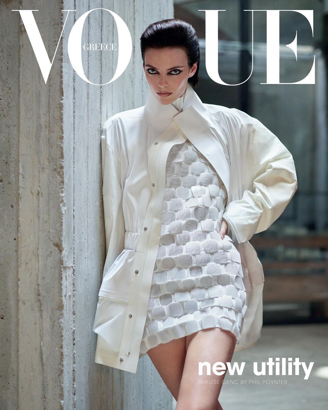 Vogue Greece cover featuring a woman in a white jacket and dress, posing against a textured concrete wall, under bright studio lighting.