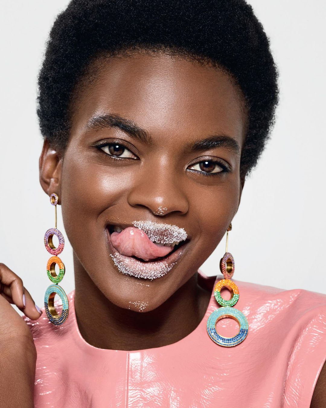 A dark-skinned woman with short afro hair and pink top is licking powdered sugar off her lips while wearing colourful circle earrings.