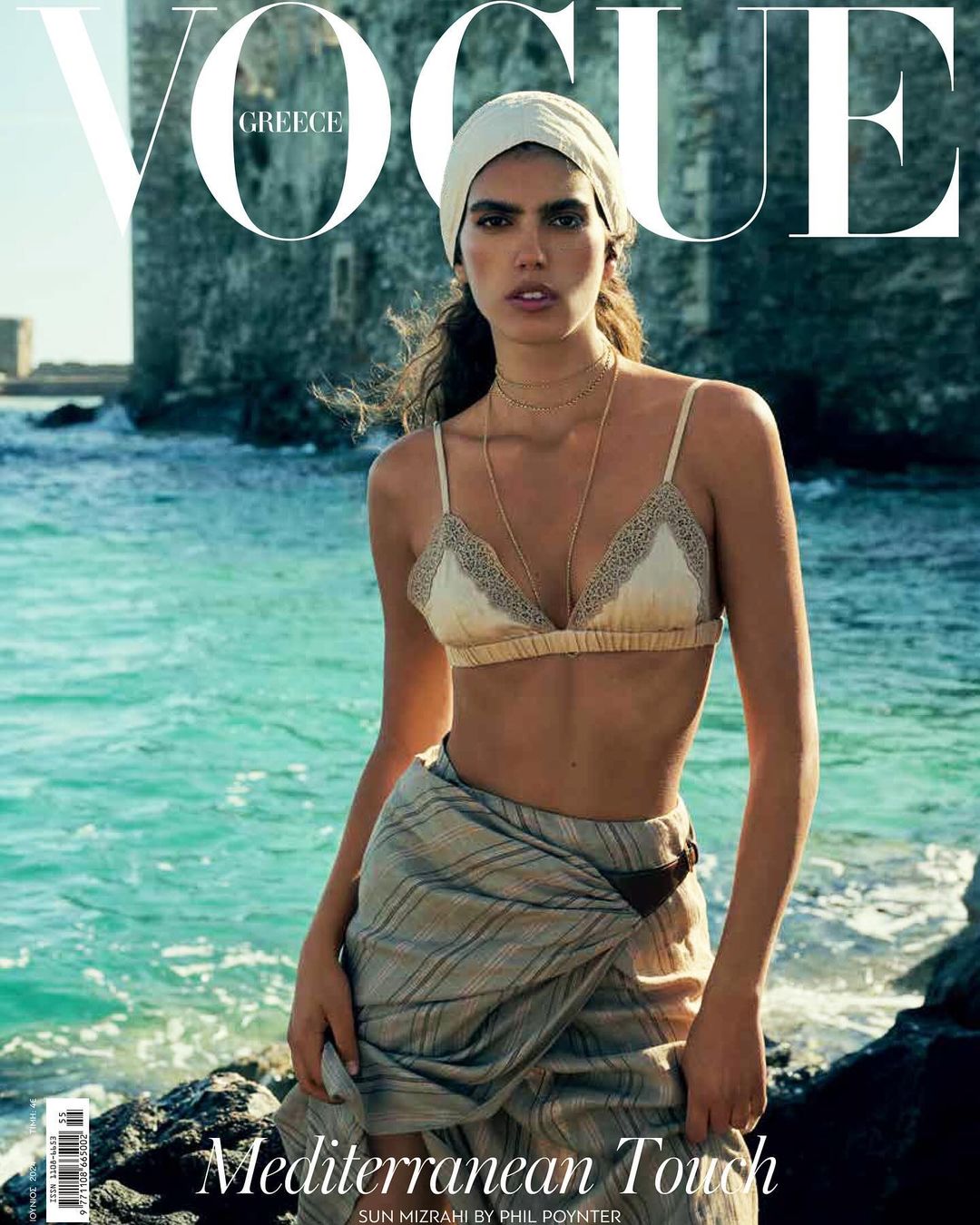 Model in a cream lace bralette and striped wrap skirt standing near rocky coastline with ocean and stone wall in background