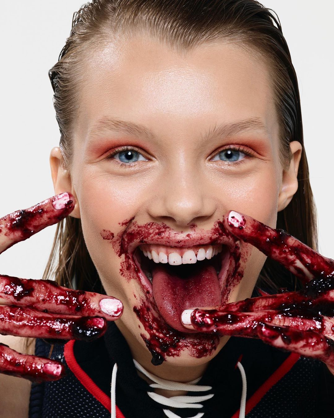 Close-up portrait of a smiling young woman with messy red makeup and dark red liquid on her face and fingers against a white background.