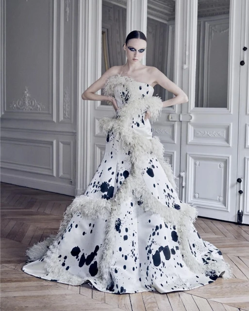 A fashion model poses indoors wearing a white, strapless gown with black abstract spots and white feather trim in front of a paneled wall.